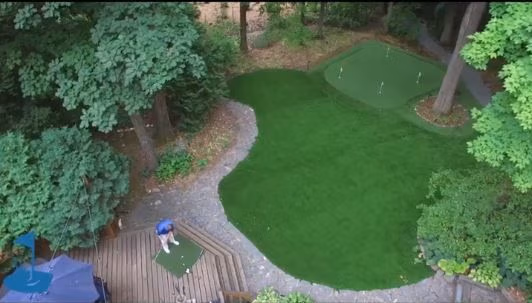 A lush, wooded backyard in Elm Grove, WI, featuring a beautifully designed artificial golf green with surrounding greenery.