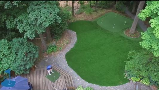 A lush, wooded backyard in Elm Grove, WI, featuring a beautifully designed artificial golf green with surrounding greenery.